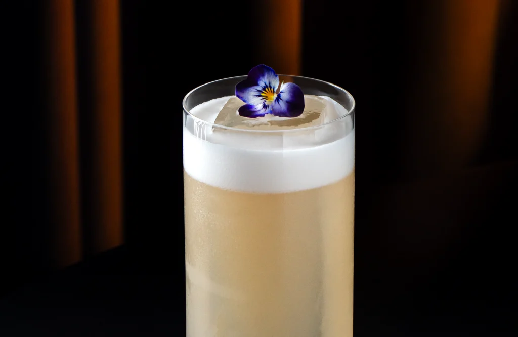 Cóctel Banana Killer de plátano con tequila reposado servido en vaso highball con espuma de aquafaba y flor comestible flotante.
